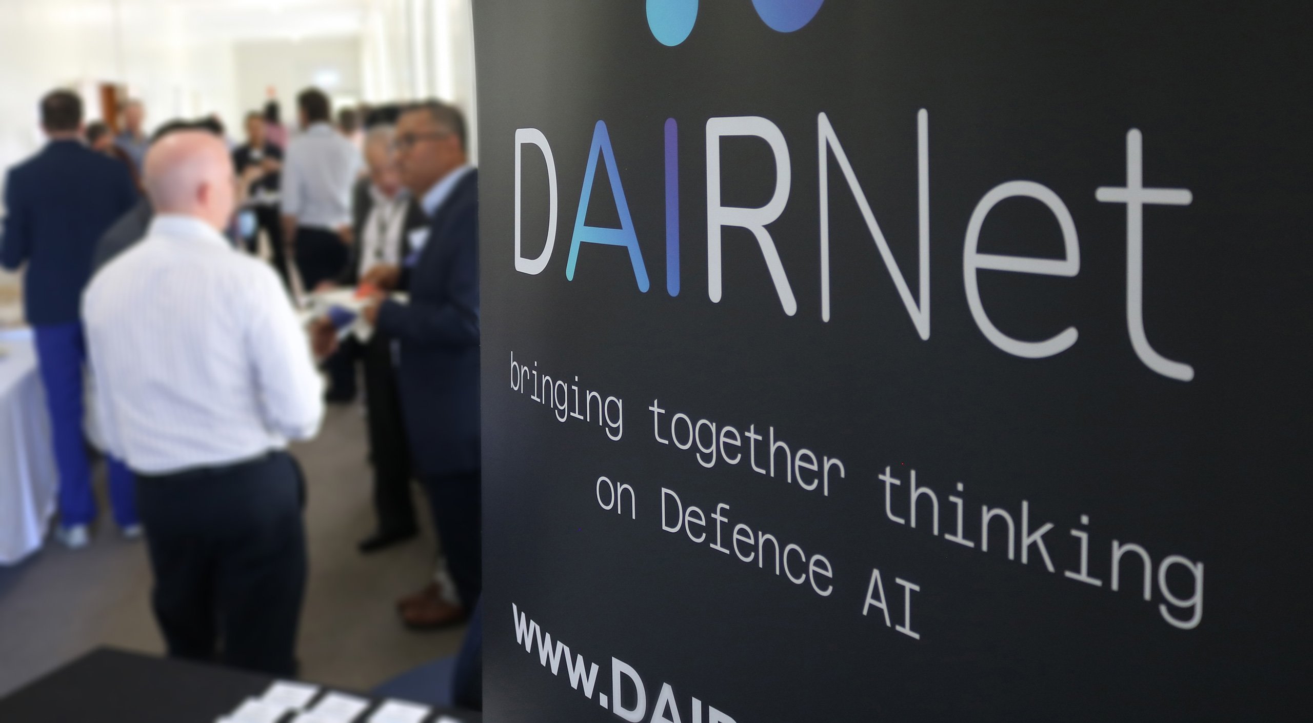 DAIRNet banner in the foreground with conference attendees talking to each other in the background.